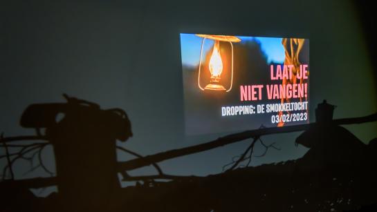 Schaduw van struikgewas en een oude telefoon in een verduisterde ruimte, met een geprojecteerde tekst: "laat je niet vangen".